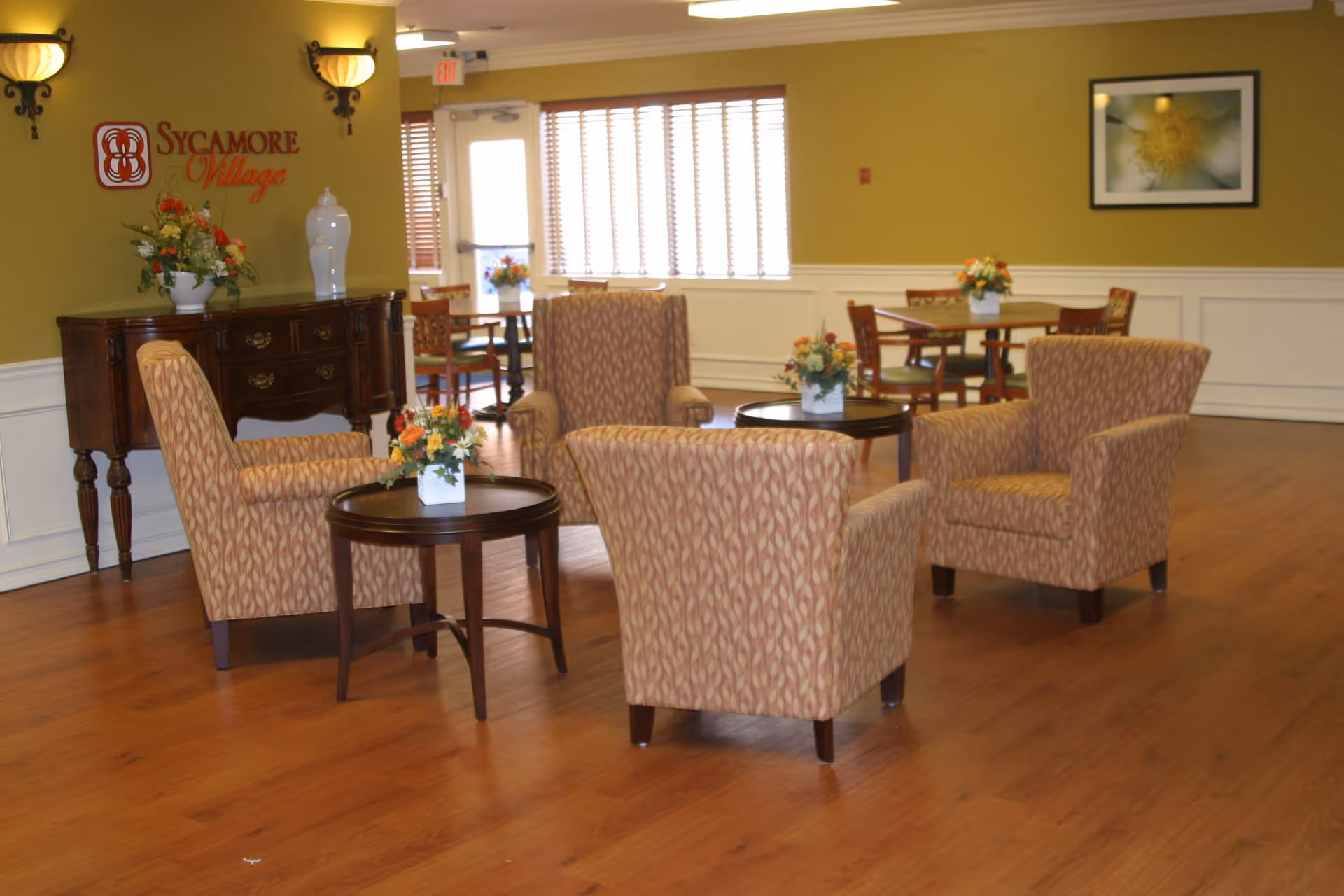 A cozy sitting area inside Sycamore Village Assisted Living featuring four patterned armchairs arranged around two small round wooden tables with floral centerpieces. The room has wooden flooring, olive green walls with white wainscoting, wall sconces, and a framed flower picture. A wooden sideboard with decorative vases and flowers is against one wall, and there are dining tables and chairs near a window with blinds in the background.