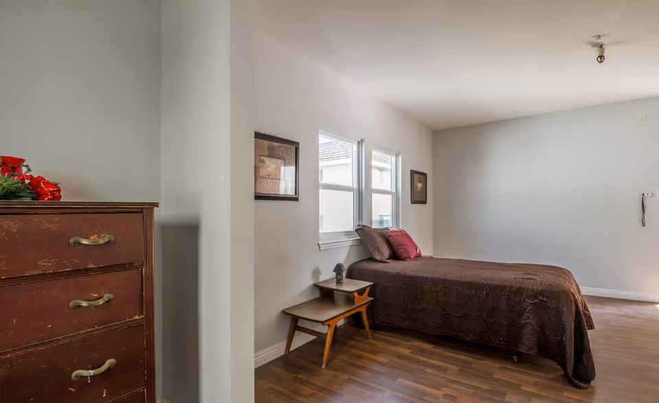 Bedroom with a single bed dressed in a brown cover, a wooden nightstand and dresser, two windows, and hardwood floors.