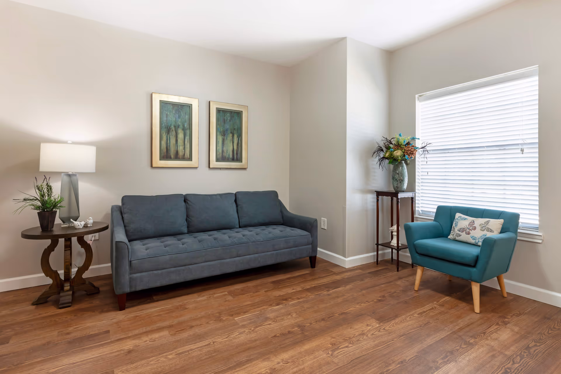 A bright living room with wooden flooring featuring a dark gray sofa with three cushions, a round wooden side table with a lamp and a small plant, two framed paintings of trees on the wall, a teal armchair with a butterfly-patterned pillow, and a small wooden stand holding a vase with flowers next to a window with white blinds.