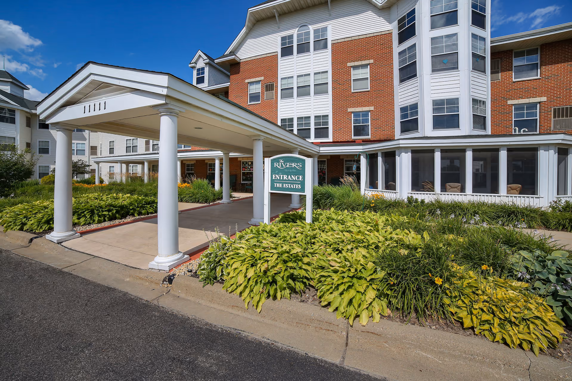 Entrance to The Rivers Retirement Community showing a covered driveway with white columns, a green sign reading 'Rivers Entrance The Estates', surrounded by well-maintained landscaping with green plants and flowers, and a multi-story brick and white building in the background under a clear blue sky.