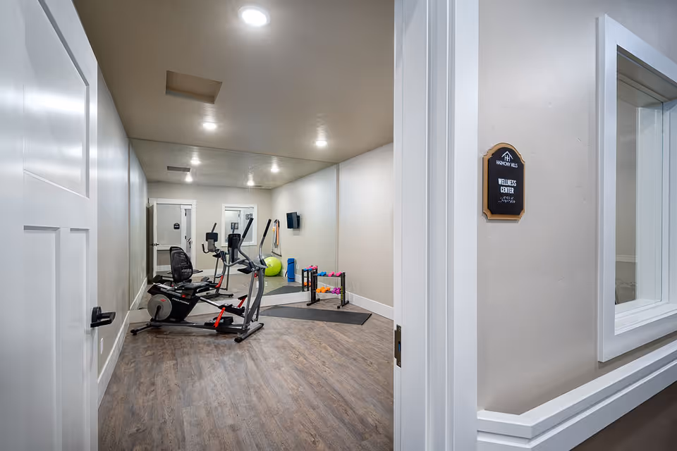 View into a wellness center room with exercise equipment including a recumbent bike, elliptical machine, yoga mats, exercise balls, and a rack of colorful dumbbells. The room has wood flooring, white walls, recessed lighting, and a large mirror on the far wall. A sign outside the room reads 'Wellness Center Harmony Hills'.