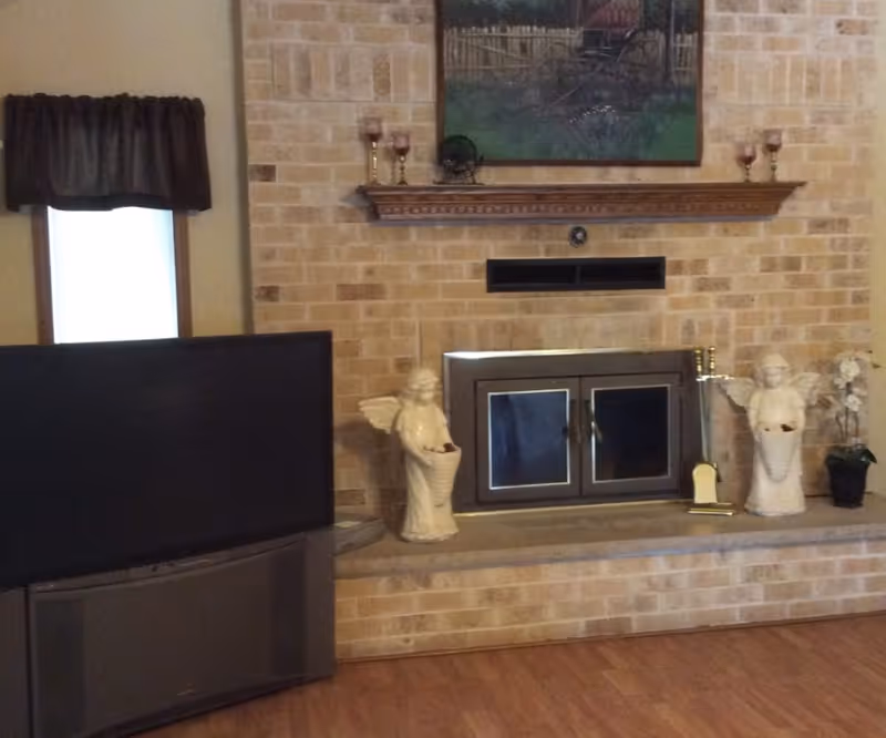 A cozy living room area featuring a brick fireplace with a wooden mantel. On the mantel are decorative candle holders and a painting of a garden scene. In front of the fireplace are two angel statues holding baskets and a small potted plant. To the left of the fireplace is a large flat-screen television on a stand, and a window with a dark curtain valance.
