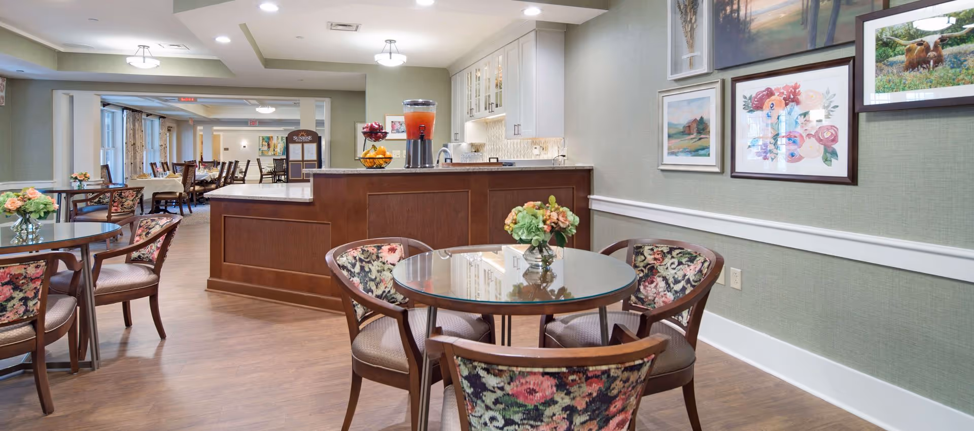 A bright and inviting dining area in a senior living facility with round glass tables and floral upholstered chairs. The room features a wooden counter with a beverage dispenser and a fruit basket. The walls are decorated with framed artwork, and the flooring is wood. In the background, there are more dining tables and chairs arranged neatly.