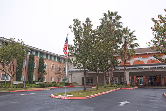 Front entrance of a multi-story senior living facility with a covered porte-cochère, circular driveway, American flag, and palm trees.