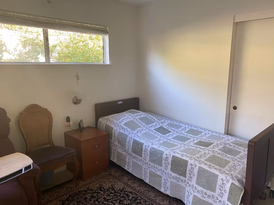 A small bedroom with a single bed covered with a patterned quilt, a wooden nightstand with two drawers, a wooden chair with a cushioned seat, a recliner chair with a tray, a window with a blind partially open, and a white wall with a small cross hanging above the nightstand.
