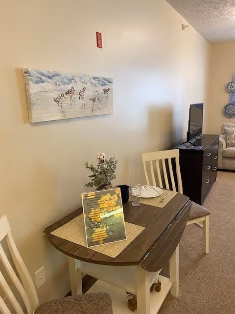 A small dining area with a wooden table set for two, featuring placemats, a glass, a plate, and a small plant centerpiece. The table is positioned against a beige wall with a painting of birds on the beach. In the background, there is a dark wooden dresser with a flat-screen TV on top and a beige couch with decorative pillows.