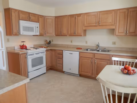 Bright kitchen with light wood cabinets, white appliances including a stove and microwave, a double sink, and a small dining table with a bowl of fruit.