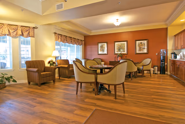 A cozy common area with wooden flooring, featuring a round table surrounded by four upholstered chairs in the center. To the left, there are two armchairs near windows with valance curtains and a small table with a lamp and flowers. The back wall is painted a warm reddish color and decorated with three framed pictures and a flower arrangement on a sideboard. On the right side, there is a kitchenette area with cabinets, a water dispenser, and coffee supplies.