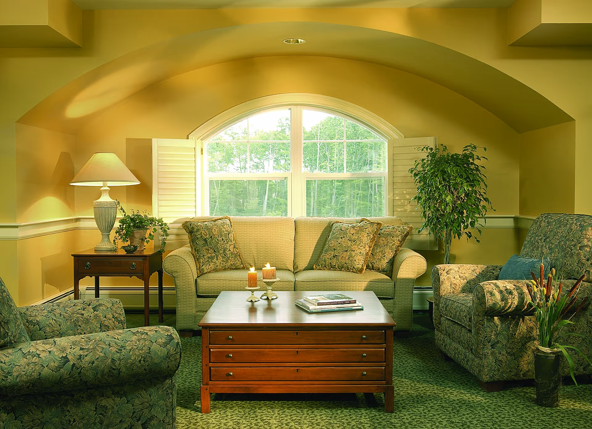 A cozy living room with a beige sofa adorned with patterned cushions, two matching armchairs, a wooden coffee table with candles and books, a side table with a lamp and plant, and a large arched window with white shutters letting in natural light. There is also a tall potted plant in the corner.