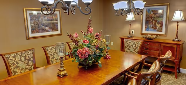 A dining room with a wooden table surrounded by six upholstered chairs with floral patterns. The table has a large floral centerpiece and two decorative candle holders. The room features two framed landscape paintings on the walls, two table lamps on a wooden sideboard, and two chandeliers with multiple lights hanging from the ceiling.