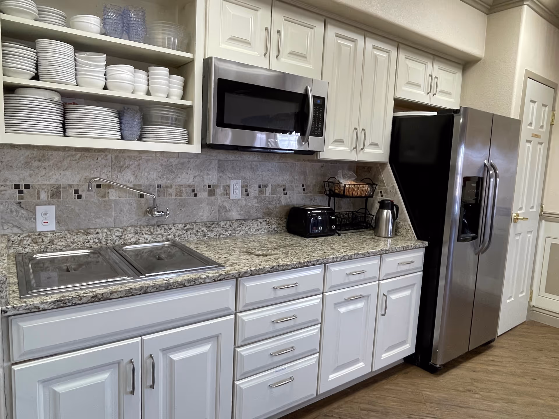 A clean and modern kitchen area with white cabinets and drawers, a granite countertop, a stainless steel microwave mounted above the counter, and a stainless steel refrigerator. Open shelves display neatly stacked white plates, bowls, and glasses. On the countertop, there is a toaster, a metal basket with bread, and a stainless steel thermal carafe. The backsplash features a tiled design with neutral tones.