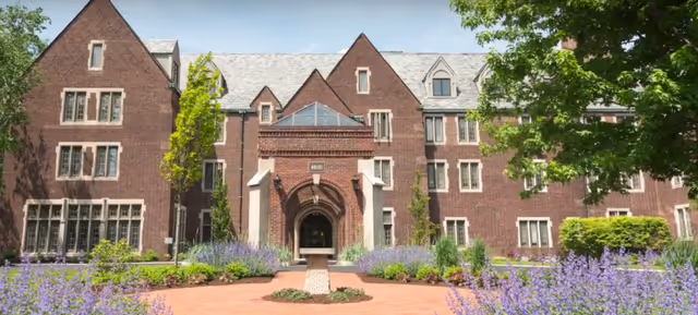Brick Tudor-style senior living building with an arched central entrance and landscaped front beds of purple flowers.