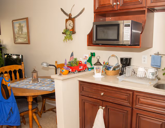 Small kitchenette and adjacent dining area with wooden cabinets, a microwave, coffee maker and dish rack on the counter, and a round table with chairs and decorative items.