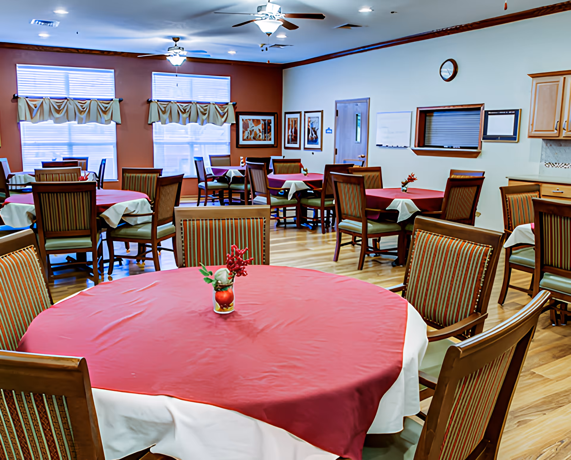 A dining room with multiple round tables covered with red and white tablecloths, each table having a small floral centerpiece. The room has wooden chairs with striped upholstery, large windows with valances, ceiling fans with lights, framed artwork on the walls, and a clock above a pass-through window to a kitchen area.