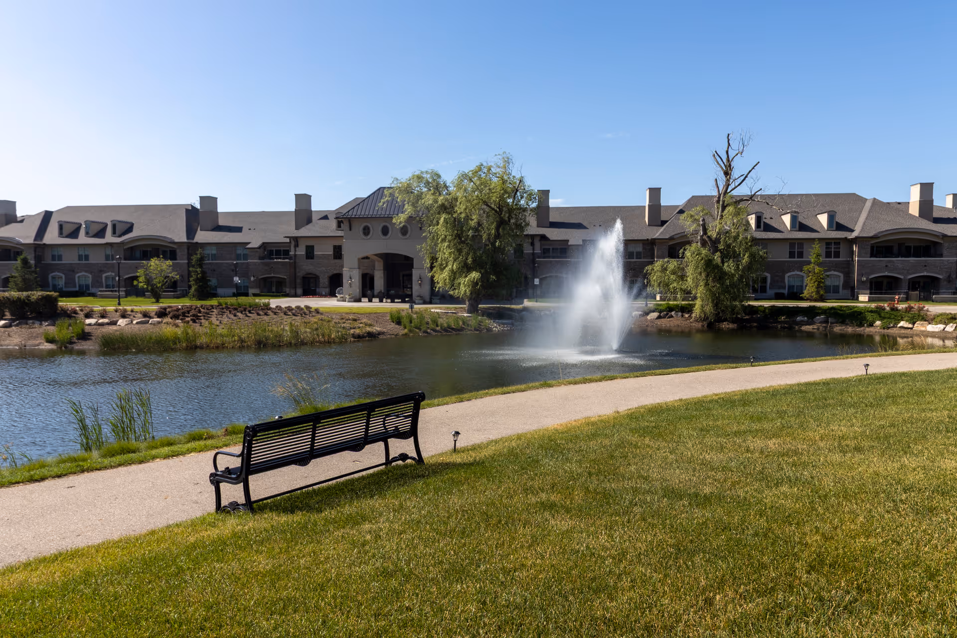 Outdoor view of a senior living facility with a large pond featuring a water fountain, a walking path, a bench, green grass, and trees under a clear blue sky.