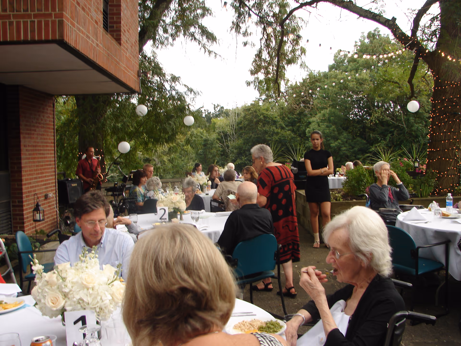 Outdoor dining event at Bethel Nursing & Rehab Center with elderly people and other guests seated at round tables covered with white tablecloths. There are floral centerpieces on the tables, string lights hanging above, and a musician playing a saxophone near a brick building. Trees and greenery surround the area.