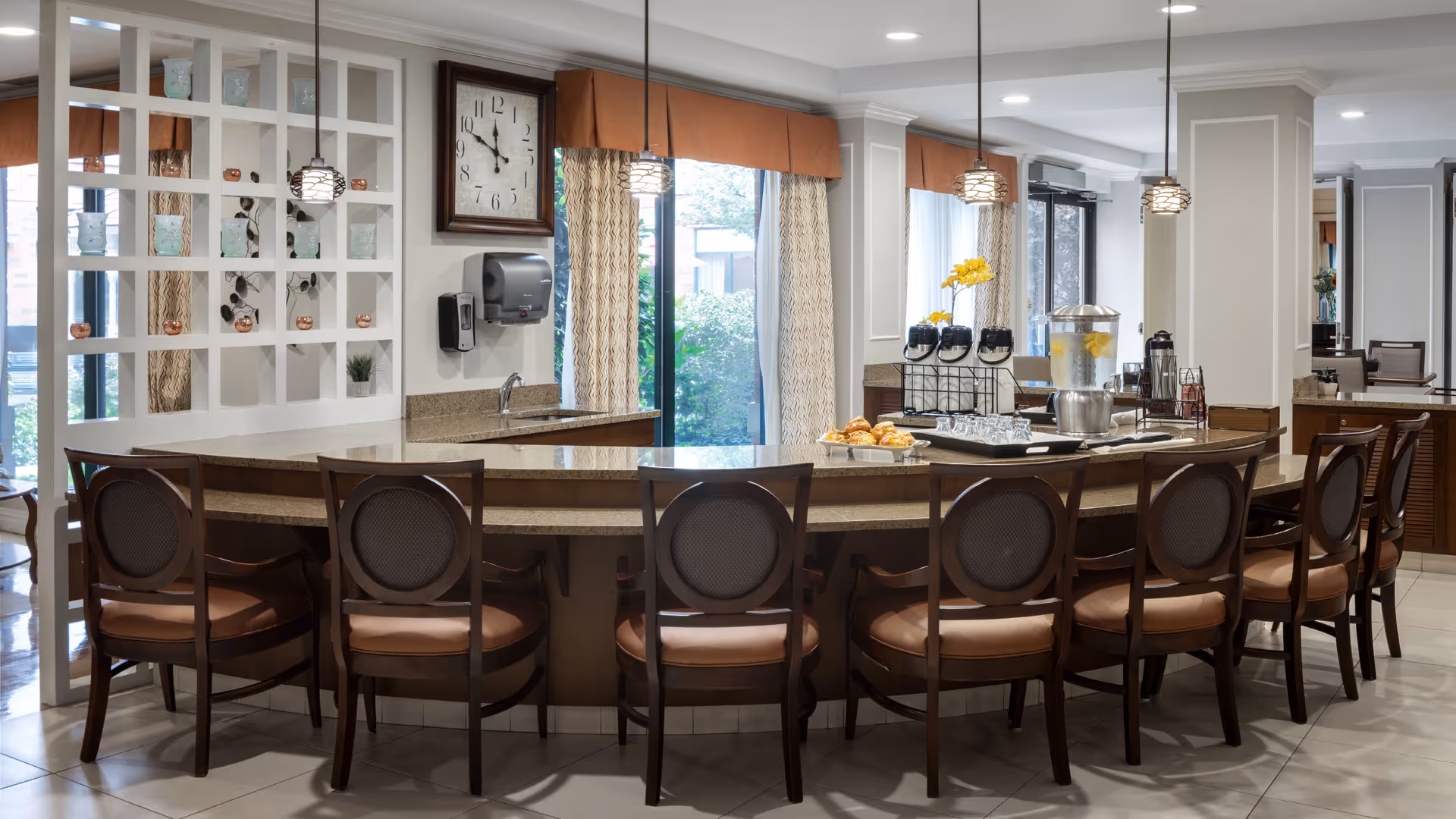 Curved counter with multiple bar stools, pendant lights, and a beverage station in a bright dining area.