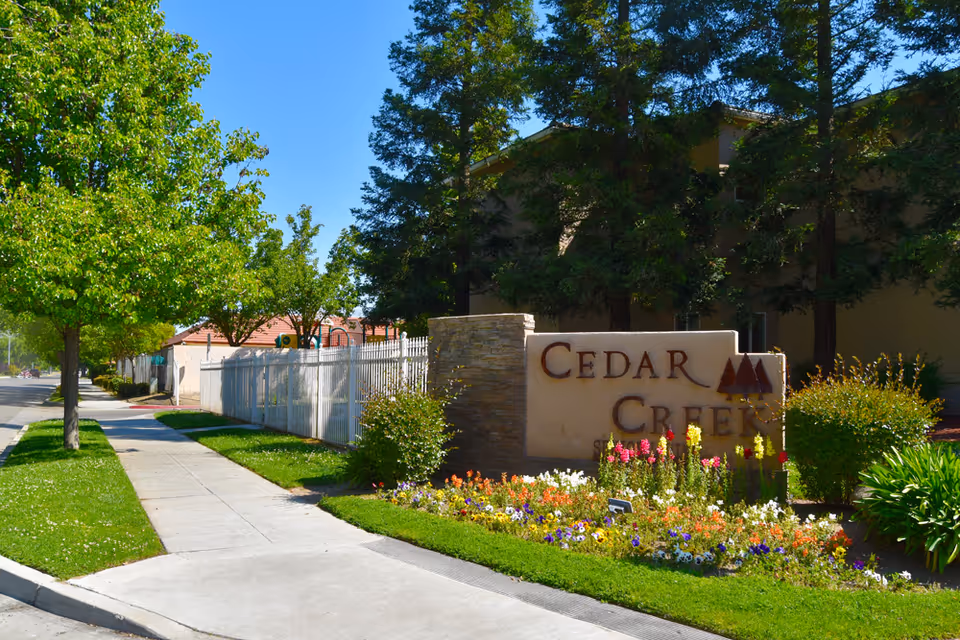 Outdoor view of the entrance area to Cedar Creek facility with a stone sign surrounded by colorful flowers and greenery, a sidewalk, trees, and a clear blue sky.