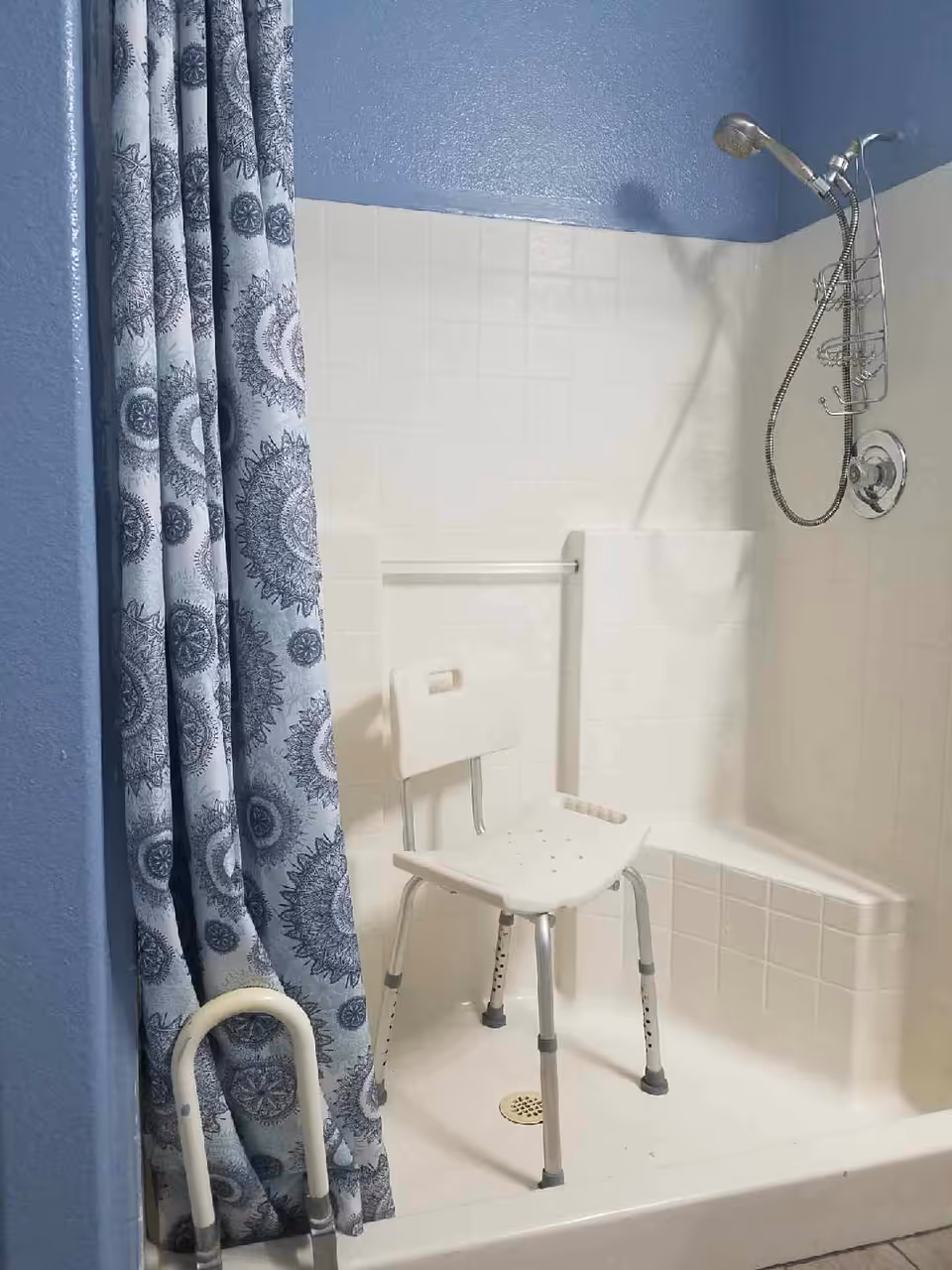 A shower area with white tiled walls and a blue painted upper wall. There is a white shower chair with metal legs inside the shower. A silver handheld showerhead is mounted on the wall with a flexible hose and a hanging shower caddy. A blue and white patterned shower curtain is partially drawn to the side. A white grab bar is visible near the entrance of the shower.