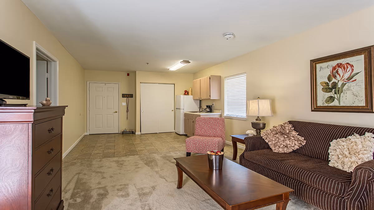 A furnished living area with a striped sofa, coffee table, patterned armchair, dresser with TV, and a small kitchenette in the background.
