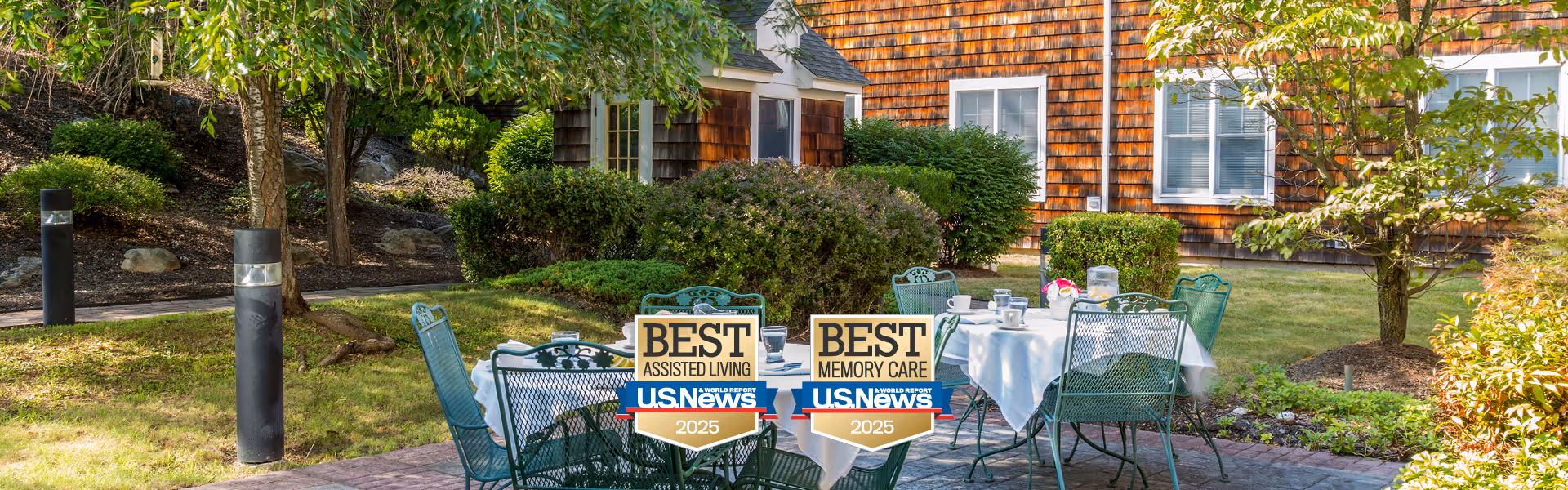 Outdoor seating area with metal chairs and tables covered with white tablecloths, set in a garden with green grass, bushes, and trees. Two signs on the chairs read 'Best Assisted Living US News 2025' and 'Best Memory Care US News 2025'. In the background, there is a building with brown shingle siding and white-framed windows.
