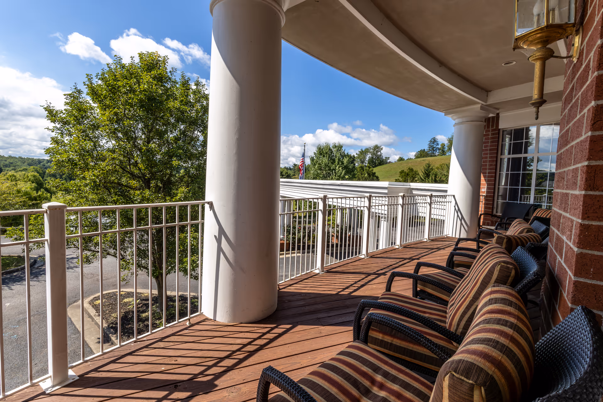 A curved outdoor balcony with wooden flooring and several cushioned chairs lined up along a brick wall. The balcony has white railings and large white columns, overlooking a green landscape with trees and hills under a partly cloudy blue sky.