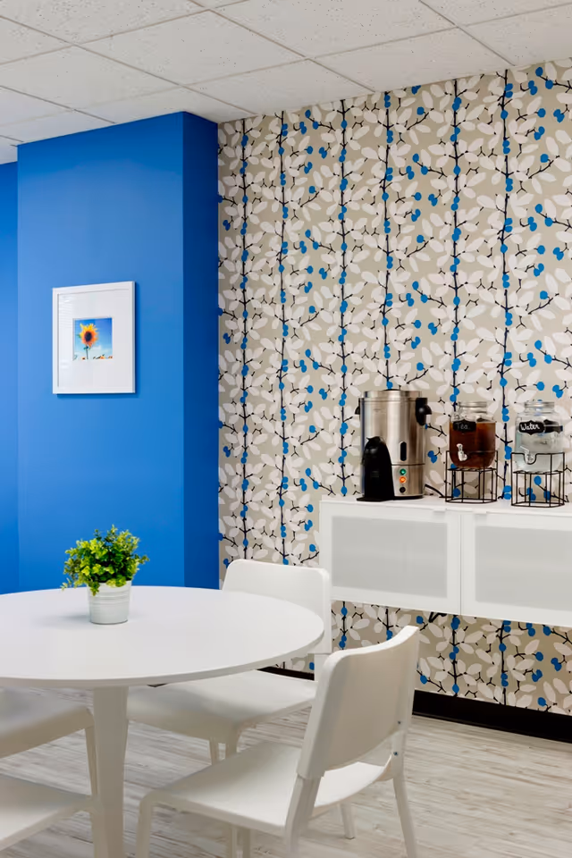 A small dining area with a round white table and four white chairs. On the table is a small green potted plant. Behind the table is a white cabinet with a coffee urn and two glass beverage dispensers labeled 'Tea' and 'Water'. The wall behind the cabinet has a patterned wallpaper with beige, white, black, and blue colors. To the left, there is a blue painted wall with a framed picture of a sunflower.