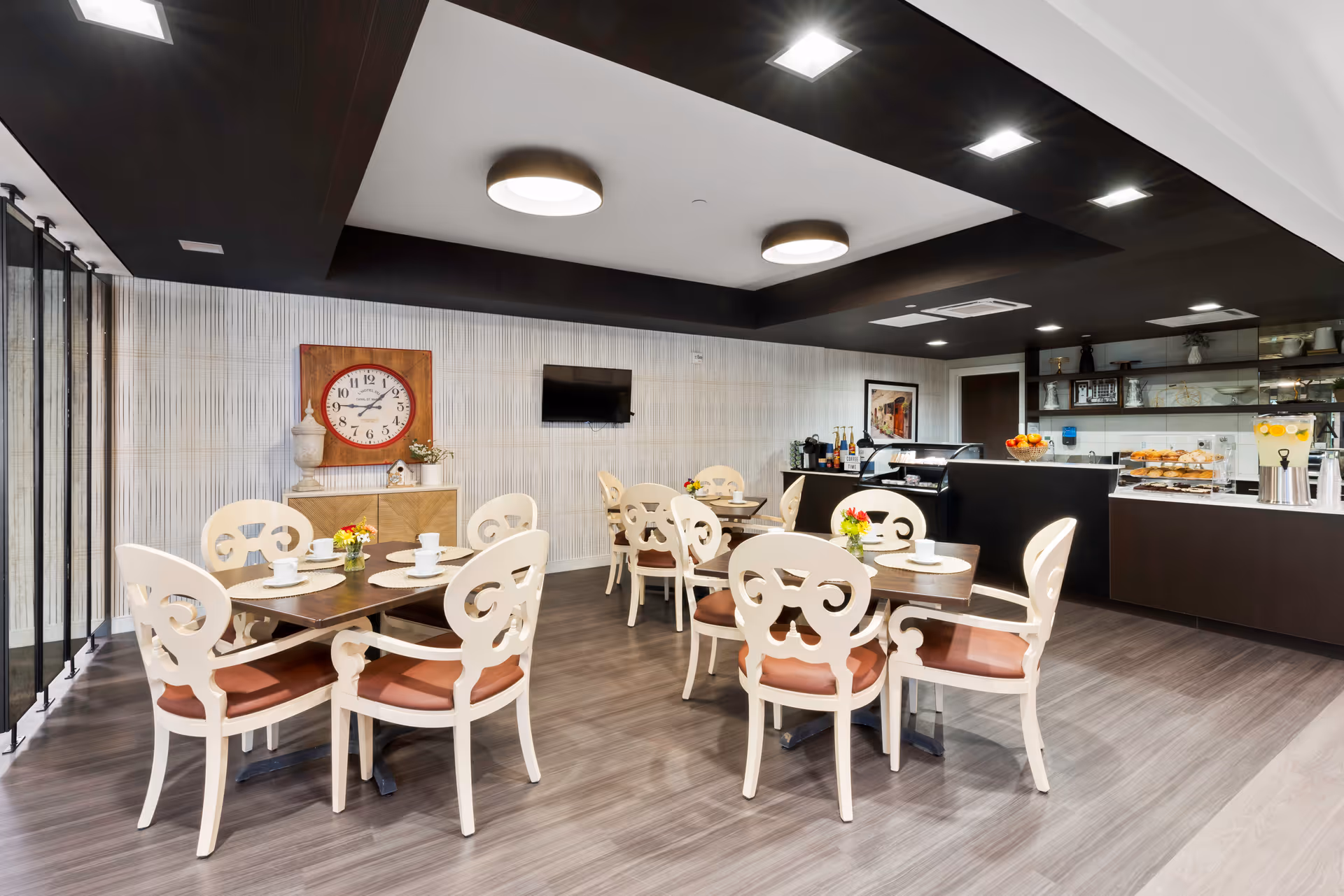 A modern dining room with several tables and white ornate chairs with brown cushions. Each table has a small flower arrangement and white cups and saucers. The room features a large wall clock, a mounted flat-screen TV, and a counter area with pastries, beverages, and a dispenser with lemonade. The ceiling has recessed lighting and circular light fixtures.