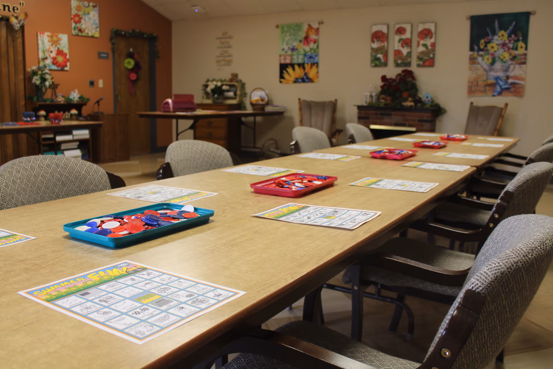 A long table in a common room set up for a game of bingo with bingo cards and trays of red, blue, and white bingo chips. The room has several chairs around the table, floral paintings on the walls, and decorative plants on tables and shelves.