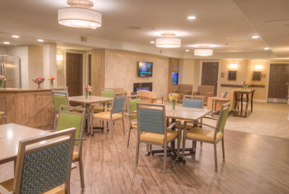 A bright and spacious common area in a senior living facility with multiple tables and chairs arranged for seating. The room features wood flooring, a fireplace with a mounted TV above it, and comfortable armchairs in the background. There are ceiling lights and vases with flowers on the tables, creating a welcoming atmosphere.