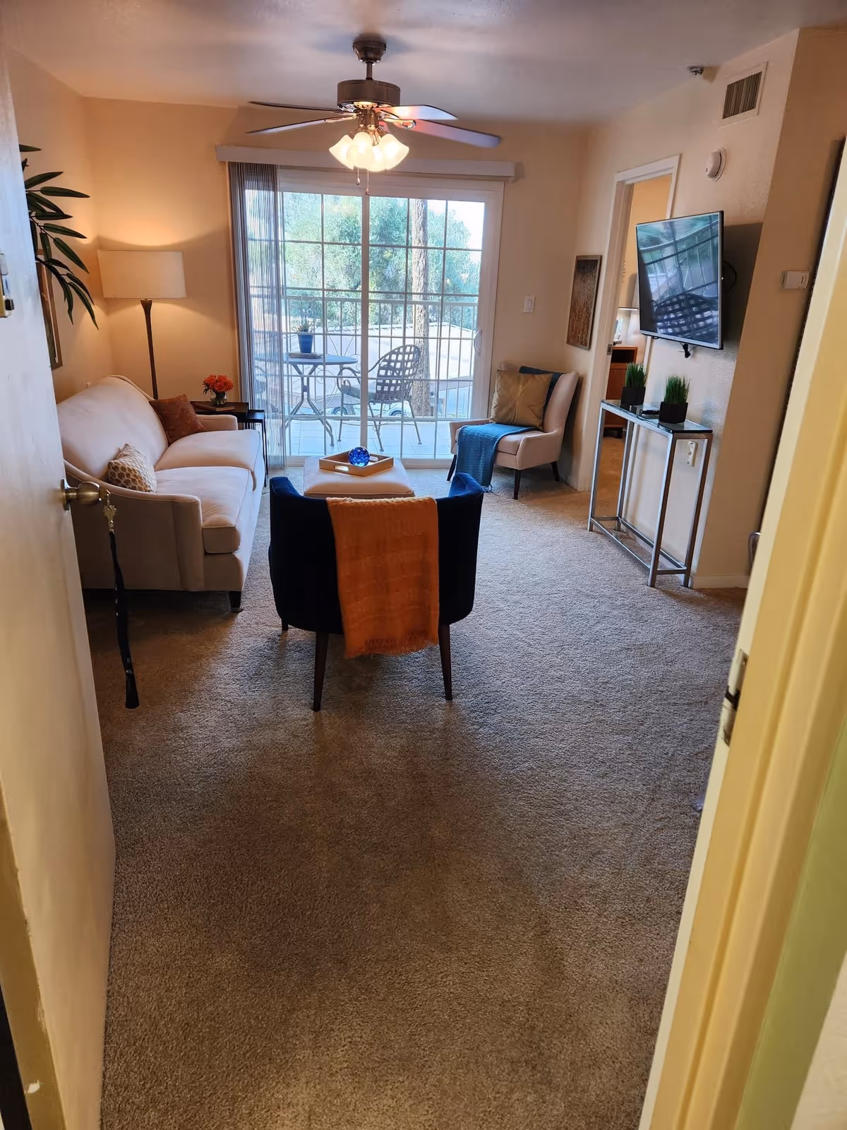A cozy living room in a senior living facility with a beige sofa, a black armchair with an orange throw blanket, a small ottoman, and a beige chair with pillows. A ceiling fan with lights is above, and a flat-screen TV is mounted on the wall. Sliding glass doors lead to an outdoor patio with a table and chairs. The room is carpeted and softly lit with a floor lamp and natural light from the patio doors.
