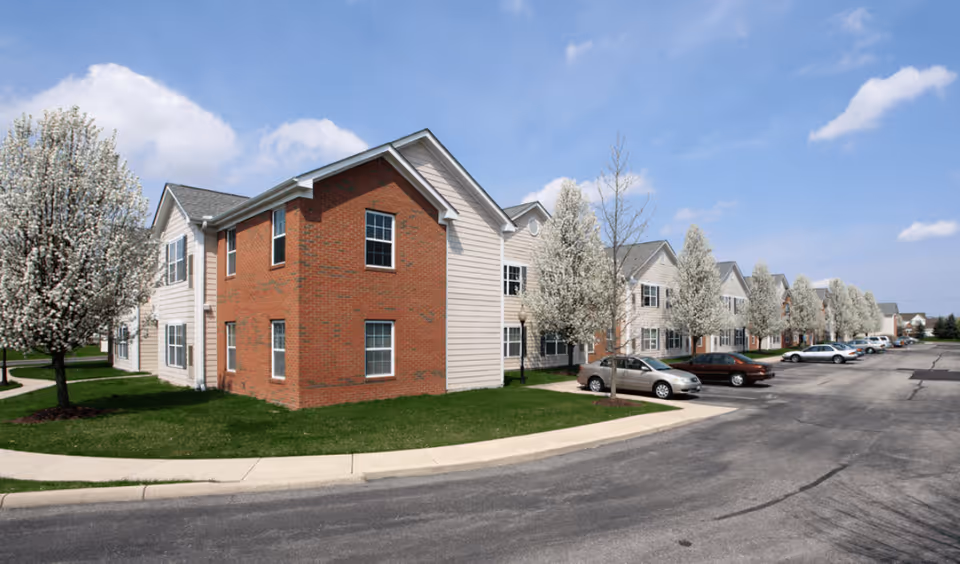 Exterior view of a two-story brick-and-siding senior apartment building with parked cars and blooming trees.