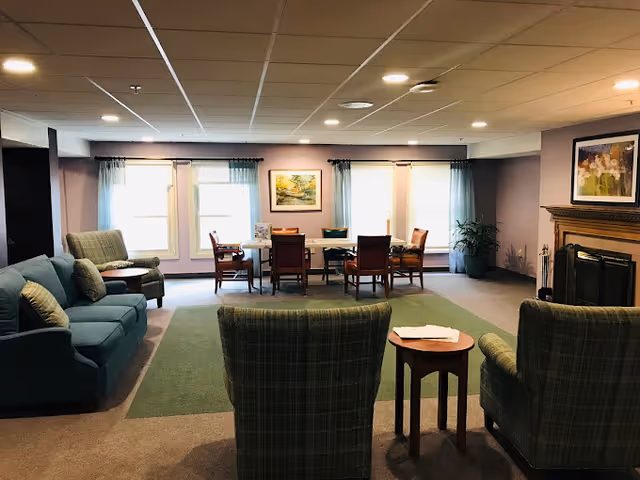 Spacious senior living common room with sofas, armchairs, a central table, windows with curtains, and a fireplace.
