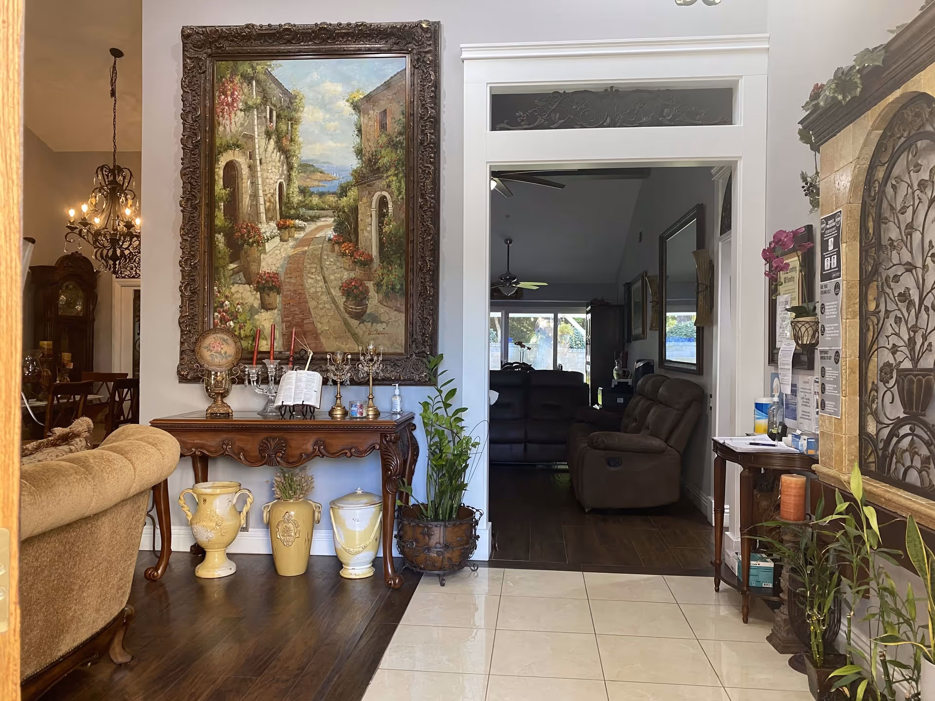 Interior view of a senior living facility showing a cozy living room area with a beige couch on the left, a wooden table with decorative vases and a large framed painting of a scenic pathway with flowers on the wall. To the right, there is a doorway leading to another room with brown recliner chairs and large windows letting in natural light. The floor transitions from dark wood to light tiles, and there are various plants and decorative items around the space.