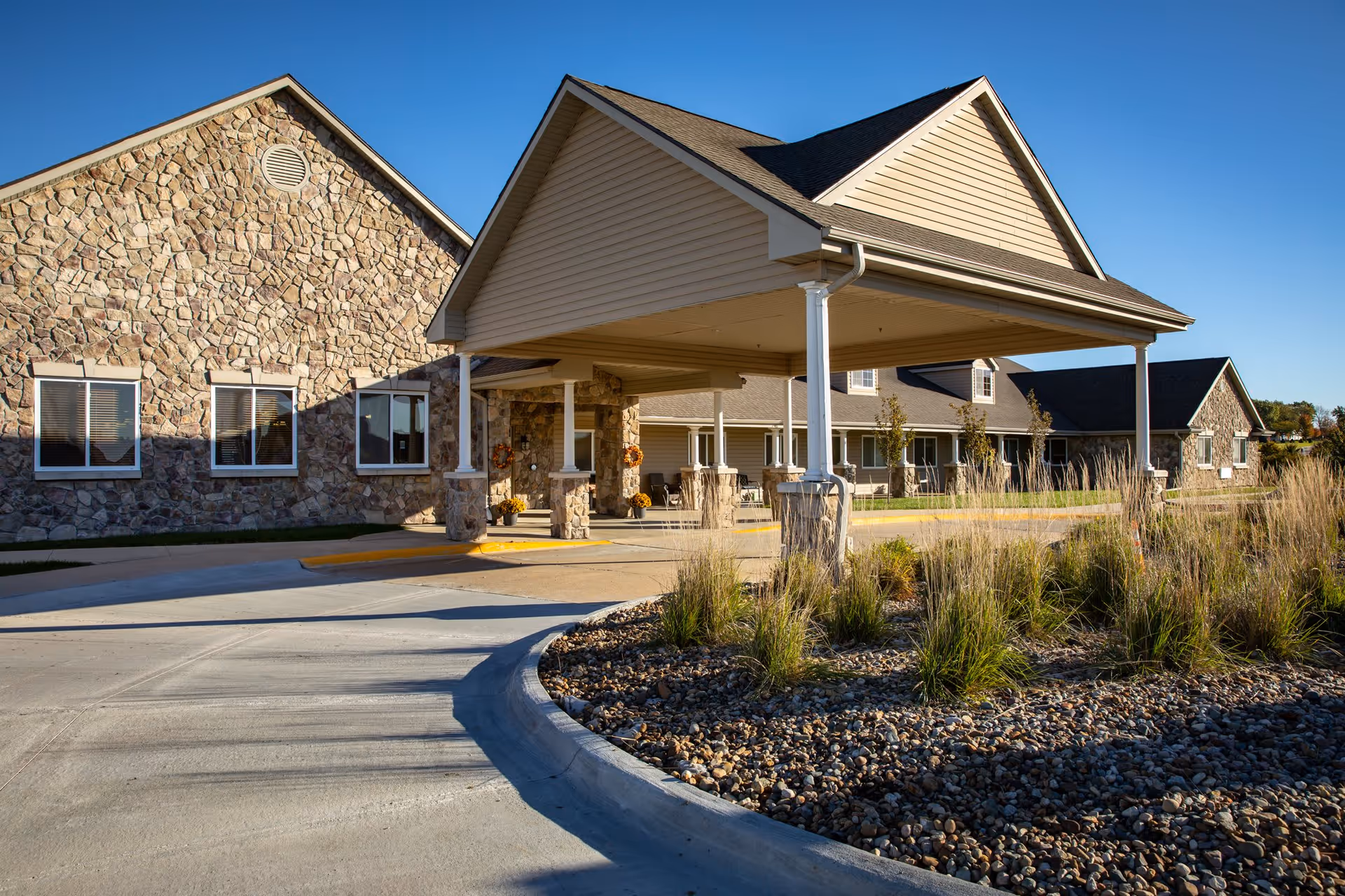 Exterior view of a senior living facility with stone and siding walls, a covered entrance supported by white columns, a curved driveway, and landscaping with rocks and tall grasses under a clear blue sky.