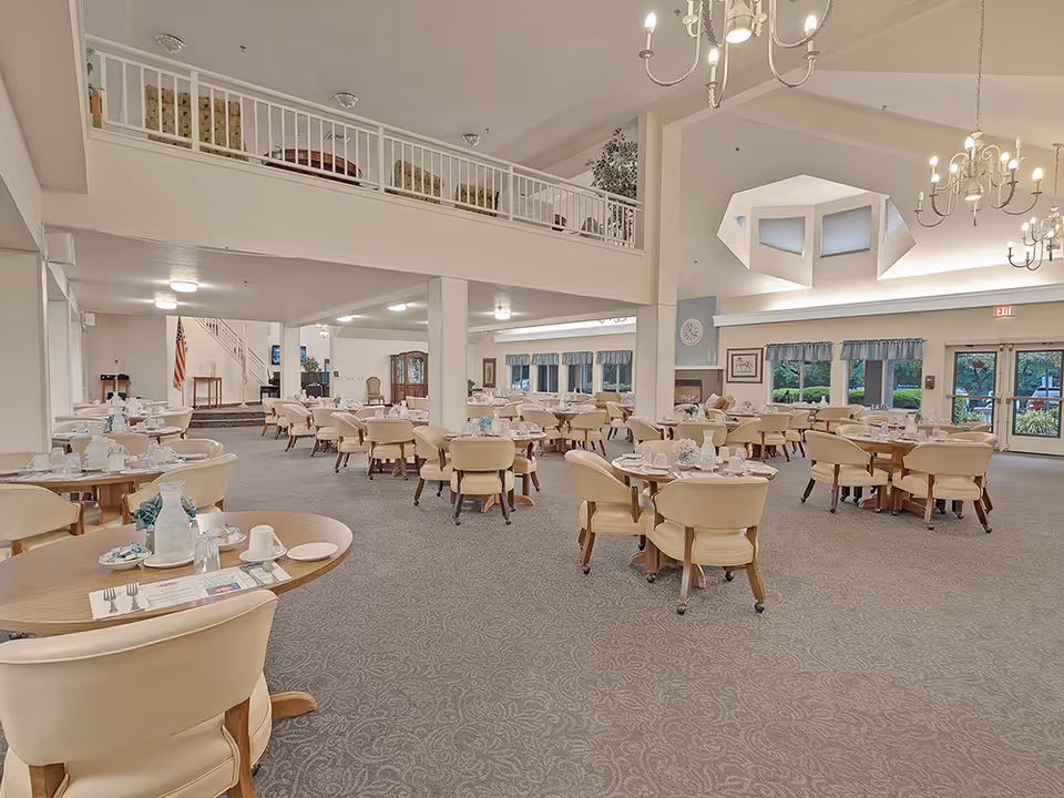 Spacious dining room in a senior living facility with multiple round tables set with white tableware and beige cushioned chairs. The room has high ceilings with chandeliers, large windows with blue valances, and a balcony area above. The carpet is patterned in a neutral tone, and there is an American flag in the background near a staircase.