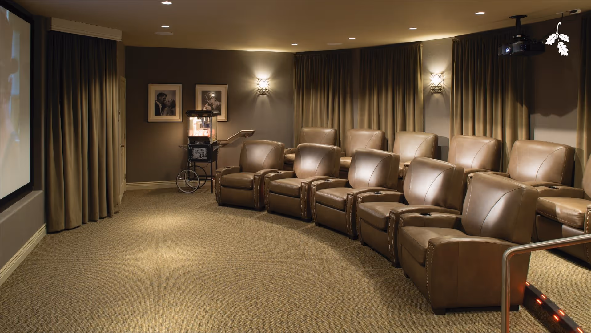 A cozy home theater room with two rows of brown leather recliner chairs facing a large projection screen. The room has beige carpet, wall sconces providing warm lighting, framed black and white photos on the wall, and a popcorn machine in the corner. Heavy curtains cover the windows, and a projector is mounted on the ceiling.