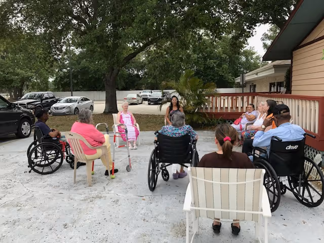 A group of elderly people sitting in a circle outdoors on a concrete surface near a building. Some are in wheelchairs, others in plastic chairs, and one uses a walker. Trees and parked cars are visible in the background.