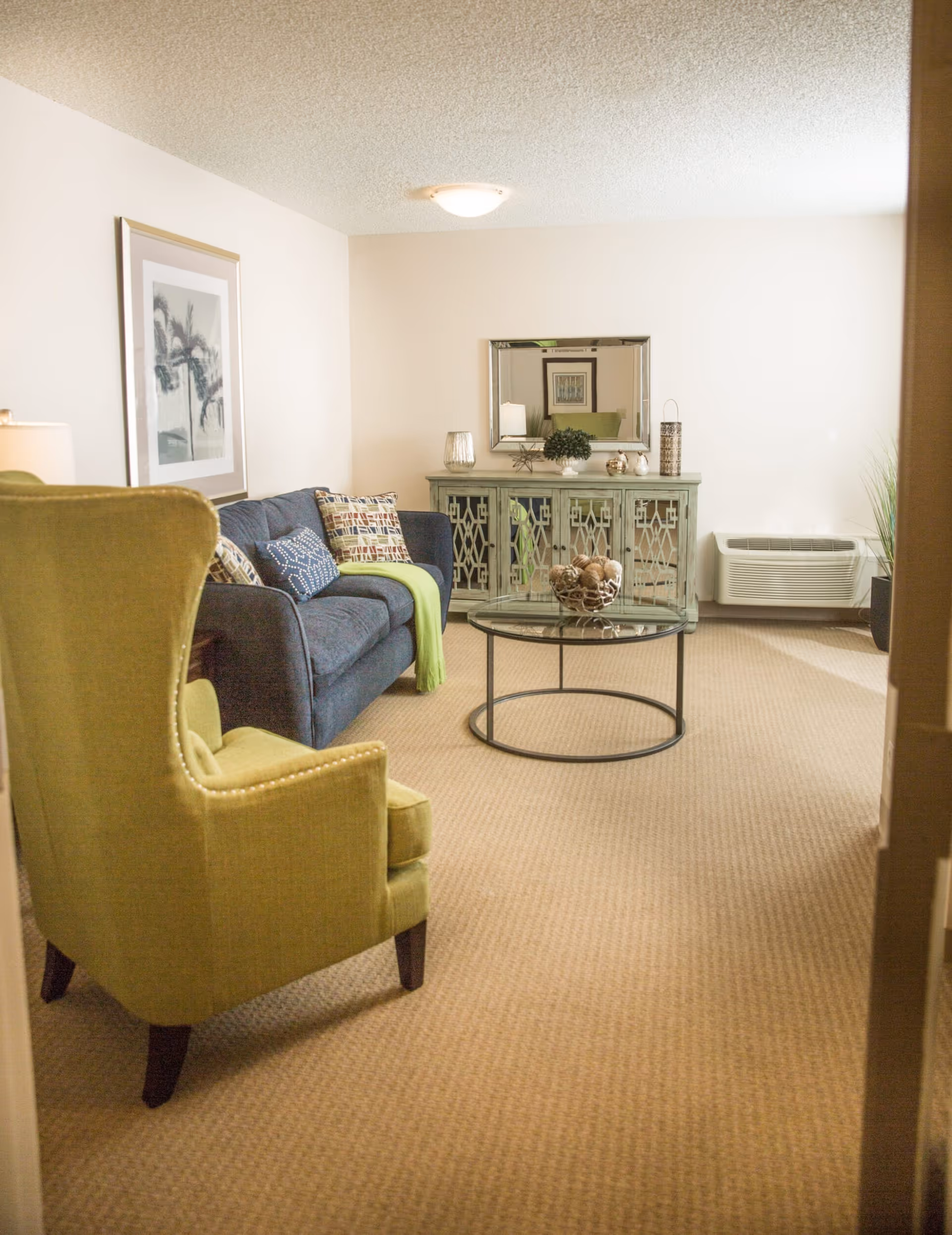 A cozy living room with a blue sofa, green armchair, round glass coffee table, and a mirrored console against a light-colored wall.