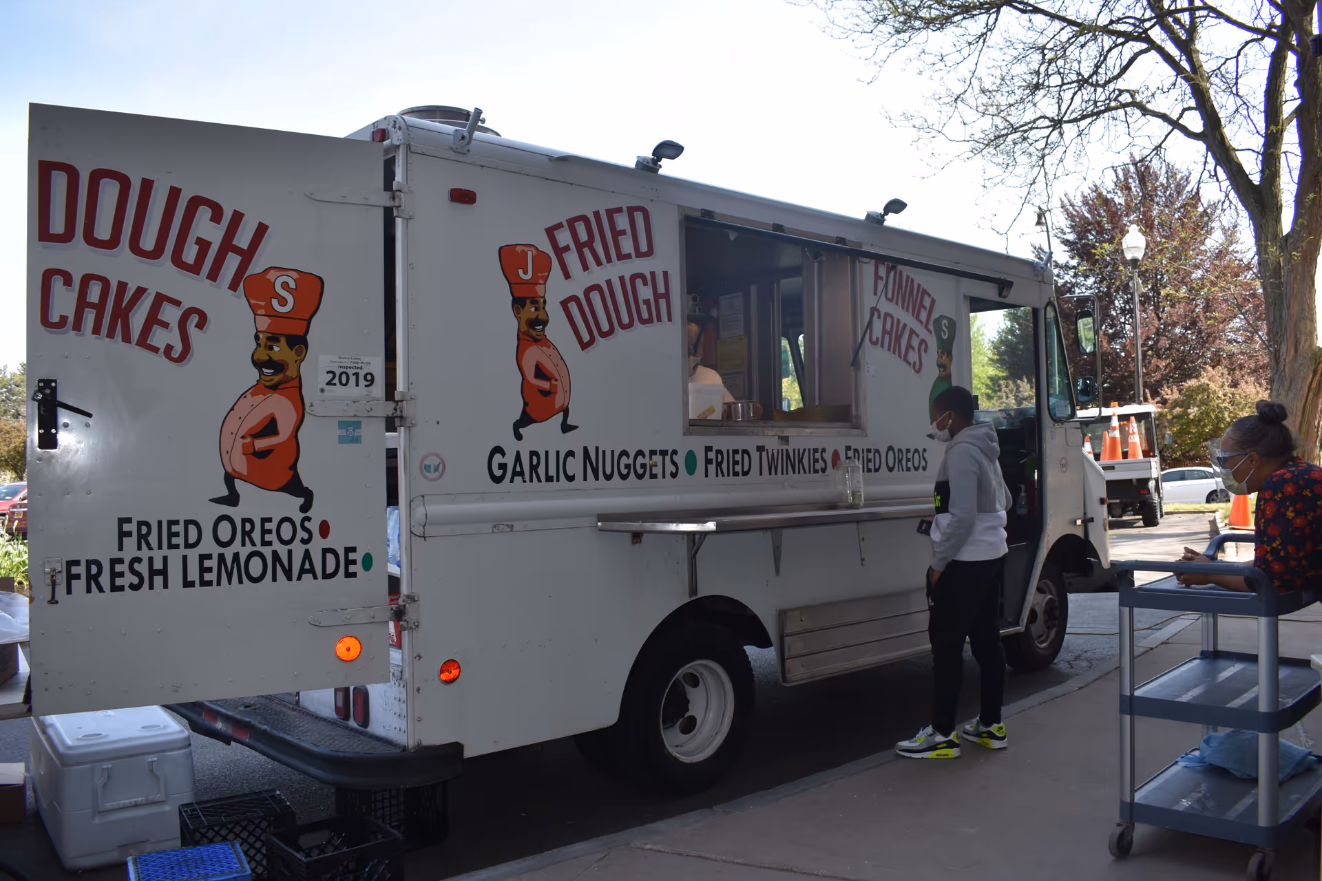 A white food truck with colorful signage advertising dough cakes, fried dough, funnel cakes, fried Oreos, garlic nuggets, fried Twinkies, and fresh lemonade. Two people wearing masks are near the truck, one standing and ordering, the other sitting on a cart. Trees and parked cars are visible in the background.