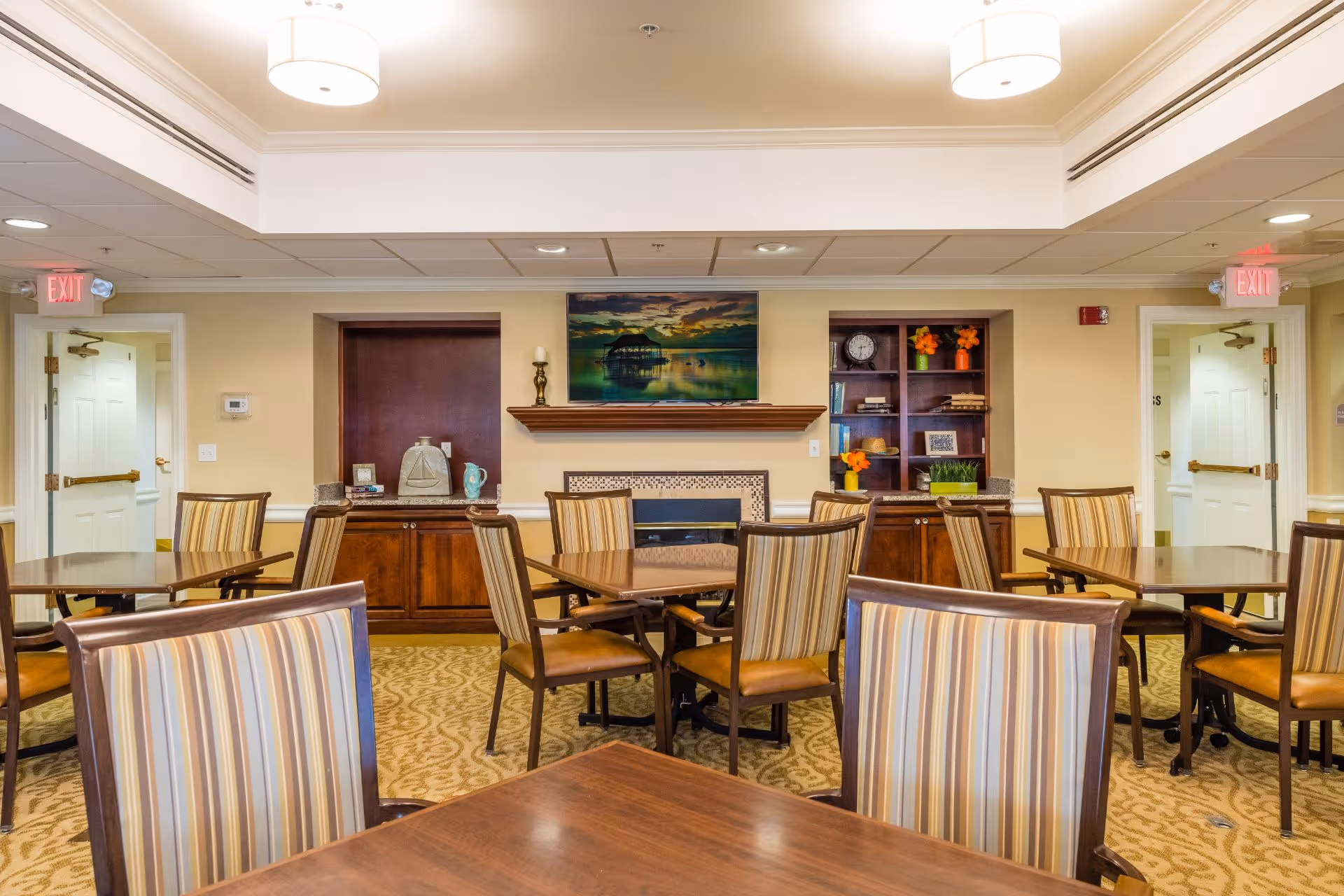 A well-lit dining room with multiple wooden tables and striped upholstered chairs arranged neatly. The room features a fireplace with a scenic painting above it, built-in wooden cabinets with decorative items, and two exit doors on either side. The ceiling has recessed lighting and two modern pendant lights.