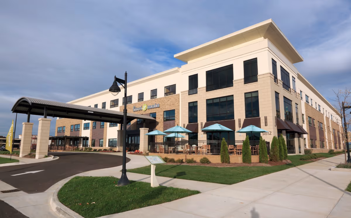 Exterior view of a modern three-story building with large windows and a covered entrance driveway. There is a patio area with tables and blue umbrellas surrounded by a black fence. The building has a mix of beige and brown brick facade with a well-maintained lawn and sidewalk in front.