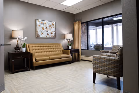A cozy sitting area in a senior living facility with a mustard yellow tufted sofa, two patterned armchairs, two dark wooden side tables with lamps and white orchids, a large window with a view of an outdoor covered patio, and a floral painting on the wall.
