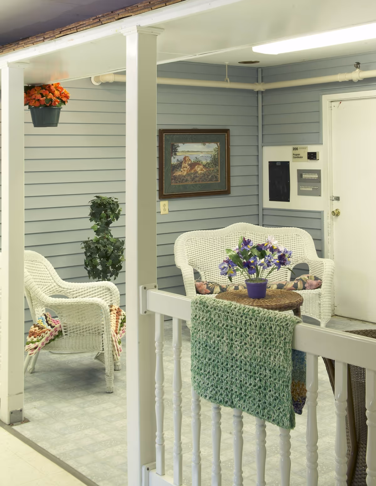 Indoor sitting area with white wicker furniture including a loveseat and a chair, both with floral cushions. A small round wicker table holds a purple pot with purple flowers. A green knitted blanket hangs over a white railing in the foreground. The walls are light blue with horizontal paneling, decorated with a framed painting of a dog lying in a natural setting. There is a white door labeled '200 Program Coordinator' and some electrical panels on the wall. A hanging pot with orange flowers and a tall green plant are also visible.
