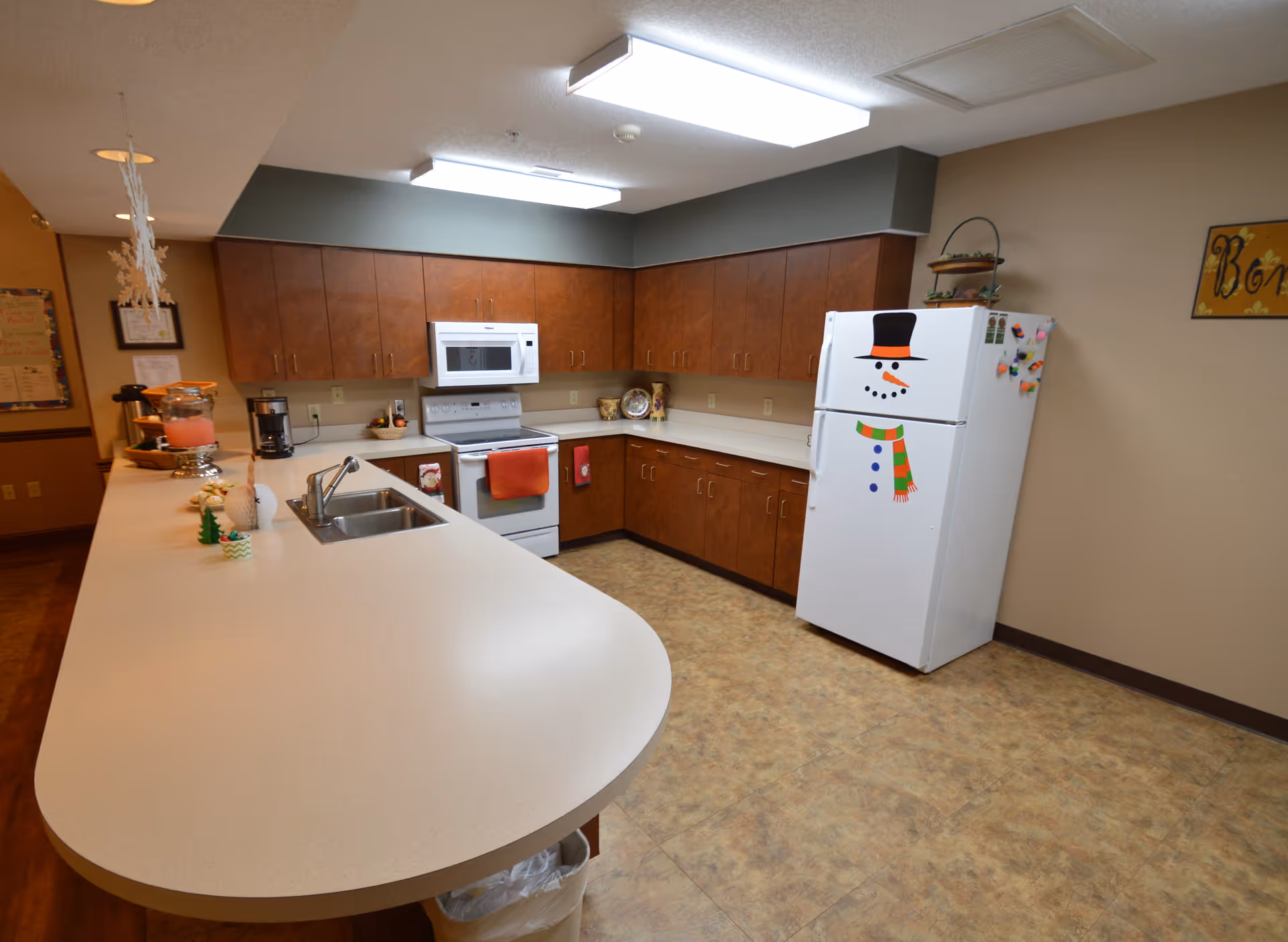 A spacious kitchen with brown wooden cabinets, a white countertop with a sink, a white stove with a microwave above it, and a white refrigerator decorated with a snowman face and scarf. The kitchen has beige walls and a tiled floor, with fluorescent ceiling lights providing illumination.