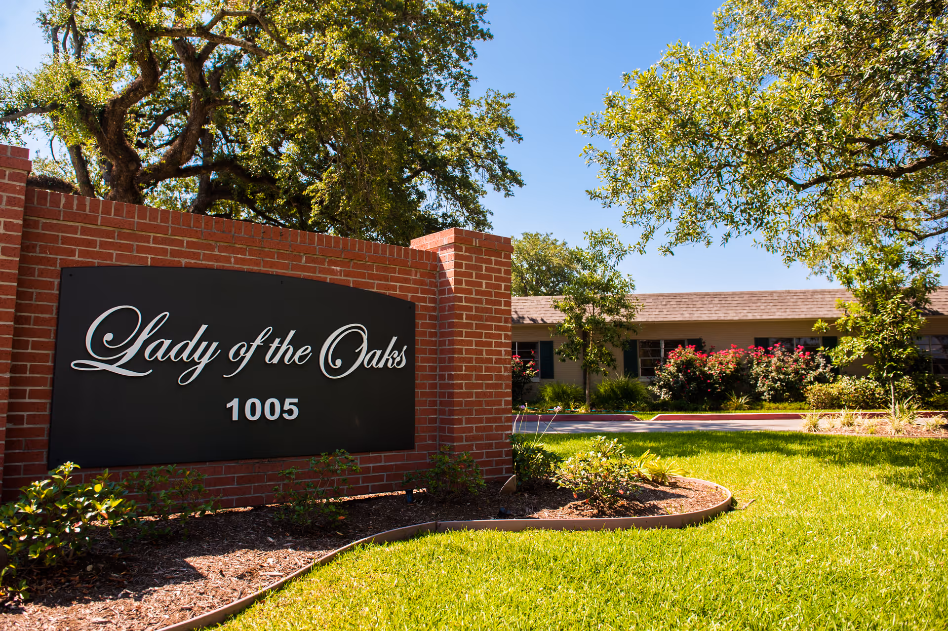 Outdoor view of the entrance sign for Lady of the Oaks Retirement facility, featuring a brick structure with a black sign displaying the name and number 1005, surrounded by green grass, bushes, and large oak trees under a clear blue sky.