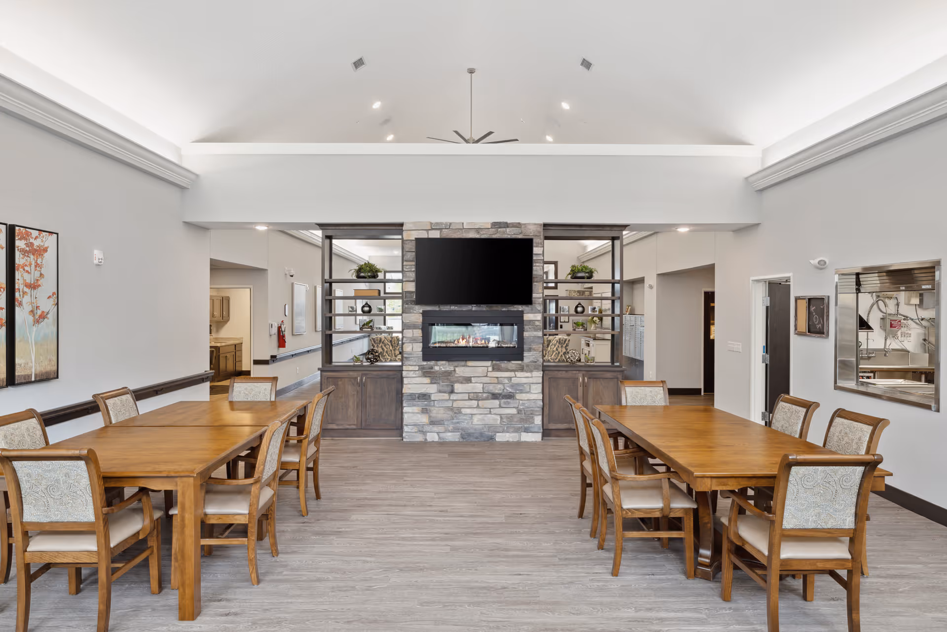 A spacious dining area in a senior living facility featuring two wooden tables each surrounded by six cushioned chairs. The room has light gray walls, a high vaulted ceiling with recessed lighting, and a central stone fireplace with a mounted flat-screen TV above it. Decorative shelving with plants and ornaments flank the fireplace. There is a pass-through window to the kitchen on the right wall and a hallway leading to other rooms on the left.