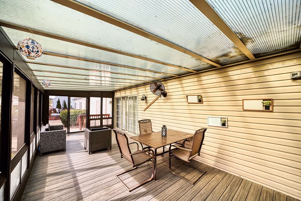 A covered outdoor patio area with a transparent roof, wooden floor, and beige siding walls. The space features a table with four chairs, a wall-mounted fan, decorative hanging lanterns, and small framed plants on the wall. There are also two cushioned armchairs near the screened windows.