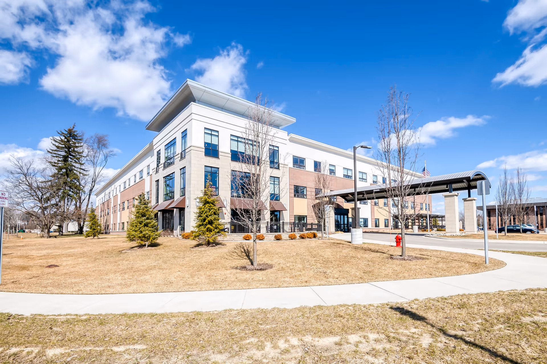 Exterior view of a modern three-story senior living facility building with large windows, a covered entrance, and a surrounding lawn with small trees under a partly cloudy blue sky.