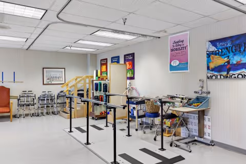 A physical therapy or rehabilitation room in a senior living facility with parallel bars for walking exercises, several walkers lined up against the wall, exercise equipment, and motivational posters on the walls.